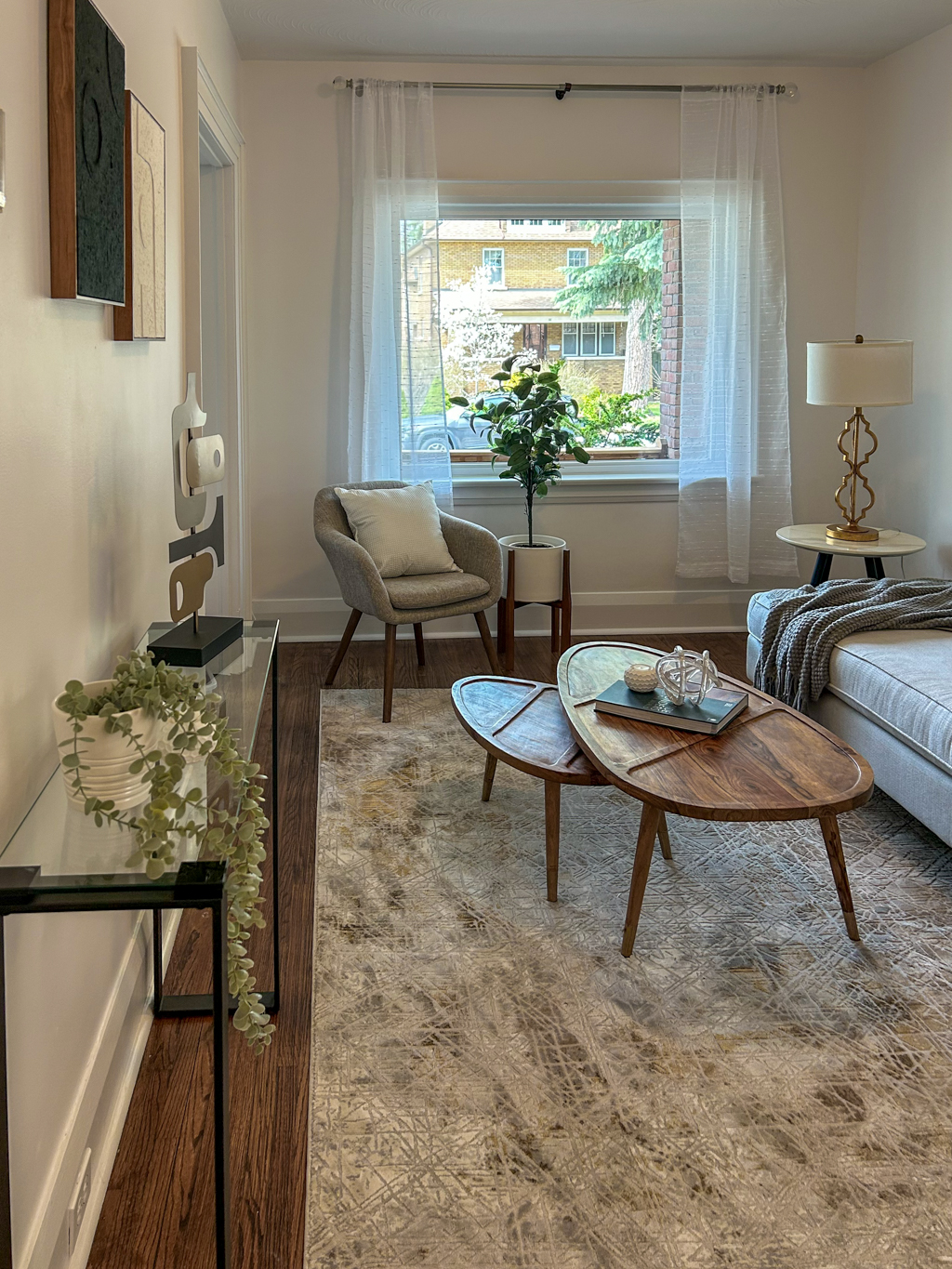 A modern, minimalistic living room in neutral tones, featuring a plush beige sofa, two sleek coffee tables, a streamlined armchair, simple side tables, abstract artwork, and a textured neutral rug. A luxury home staging project by Hermosa Home Interior.